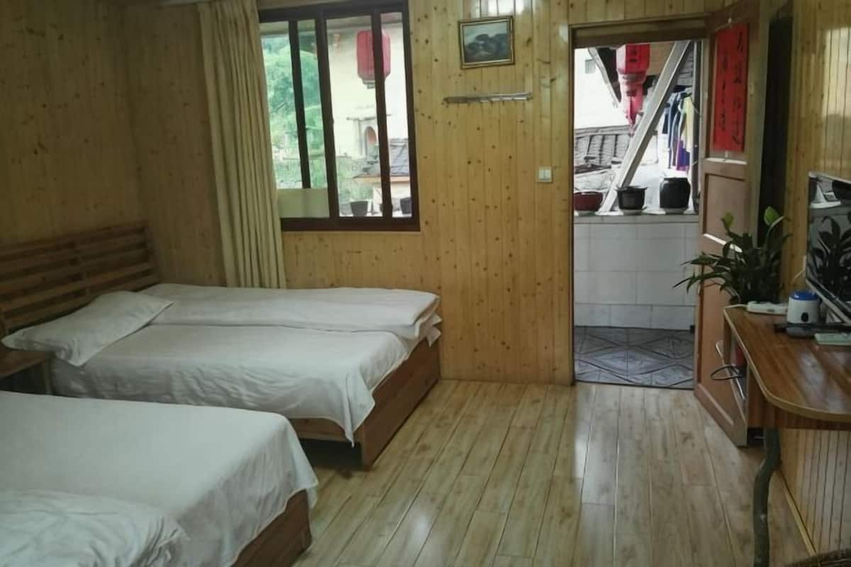 A room with two beds, a wooden floor, a window with curtains, a small table with a plant, and a view of a staircase.