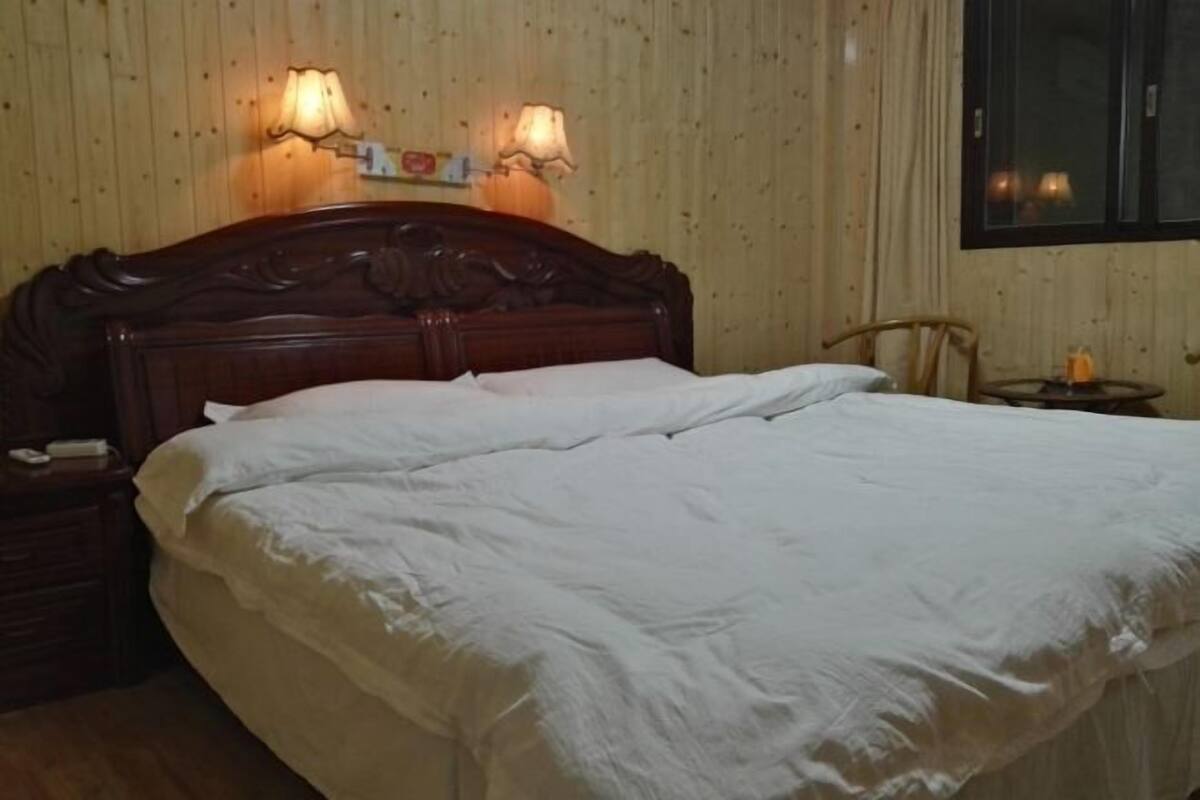 A neatly made bed in a room with wooden walls and a window. There are two wall-mounted lamps and a small table with a candle.