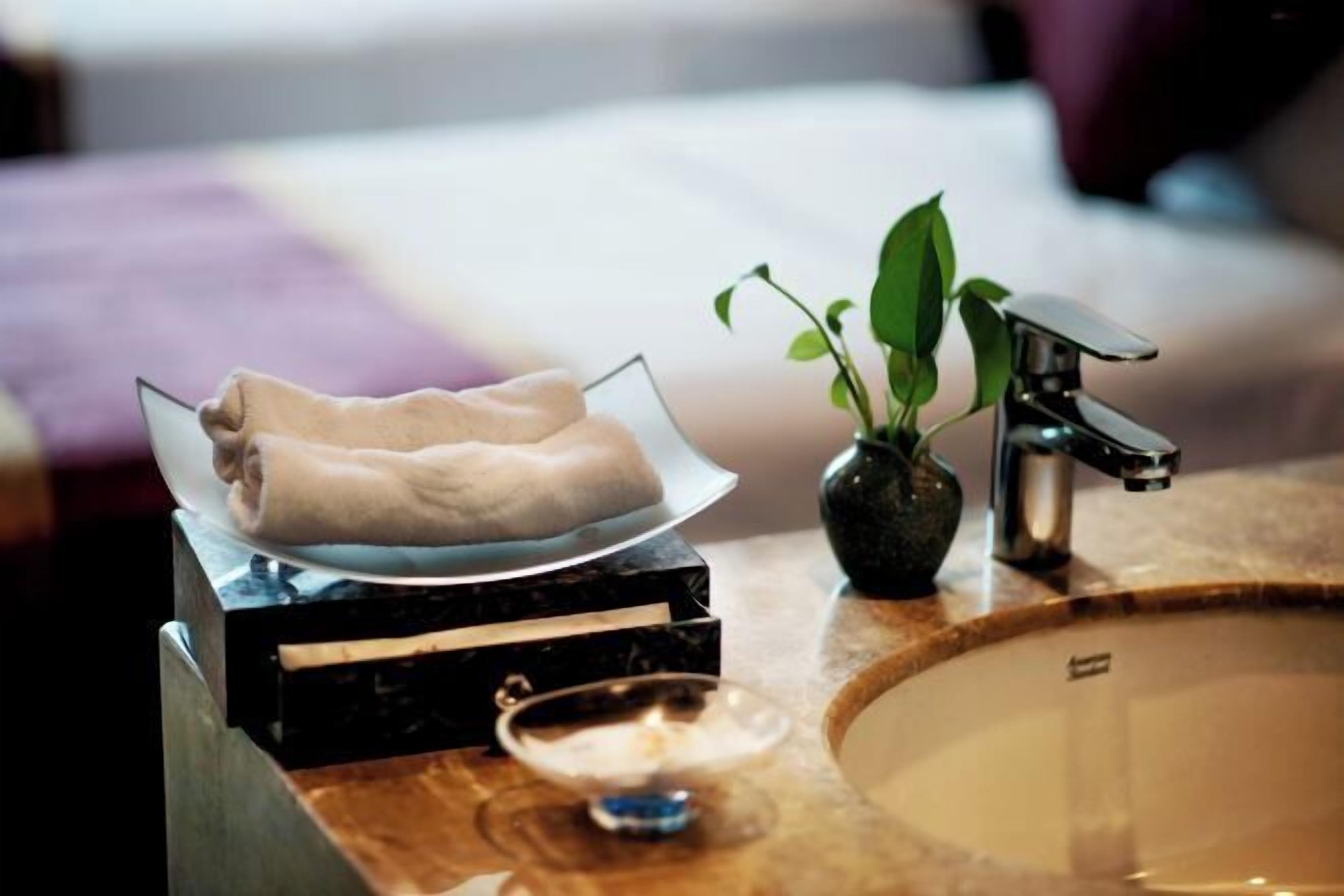 A bathroom with a marble sink, a glass bowl with towels, a potted plant, and a modern faucet.