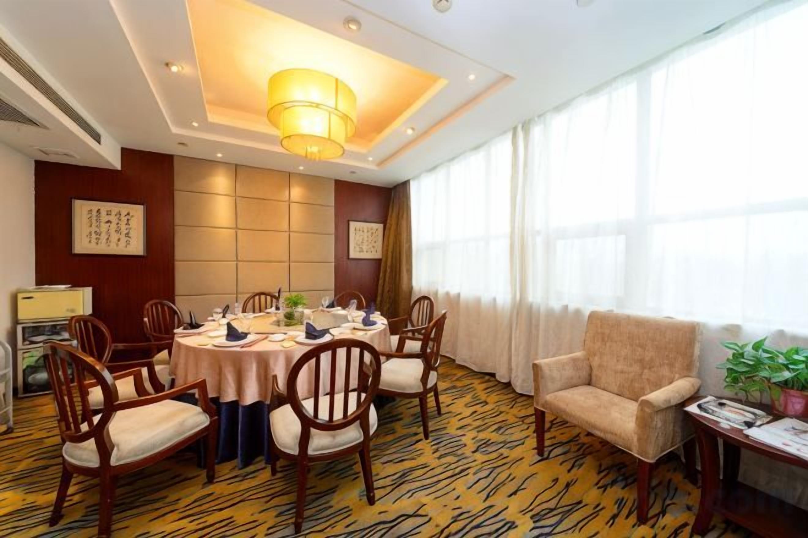 A hotel dining room with a round table, chairs, a sofa, and a large window with curtains.