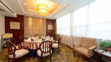 A hotel dining room with a round table, chairs, a sofa, and a large window with curtains.