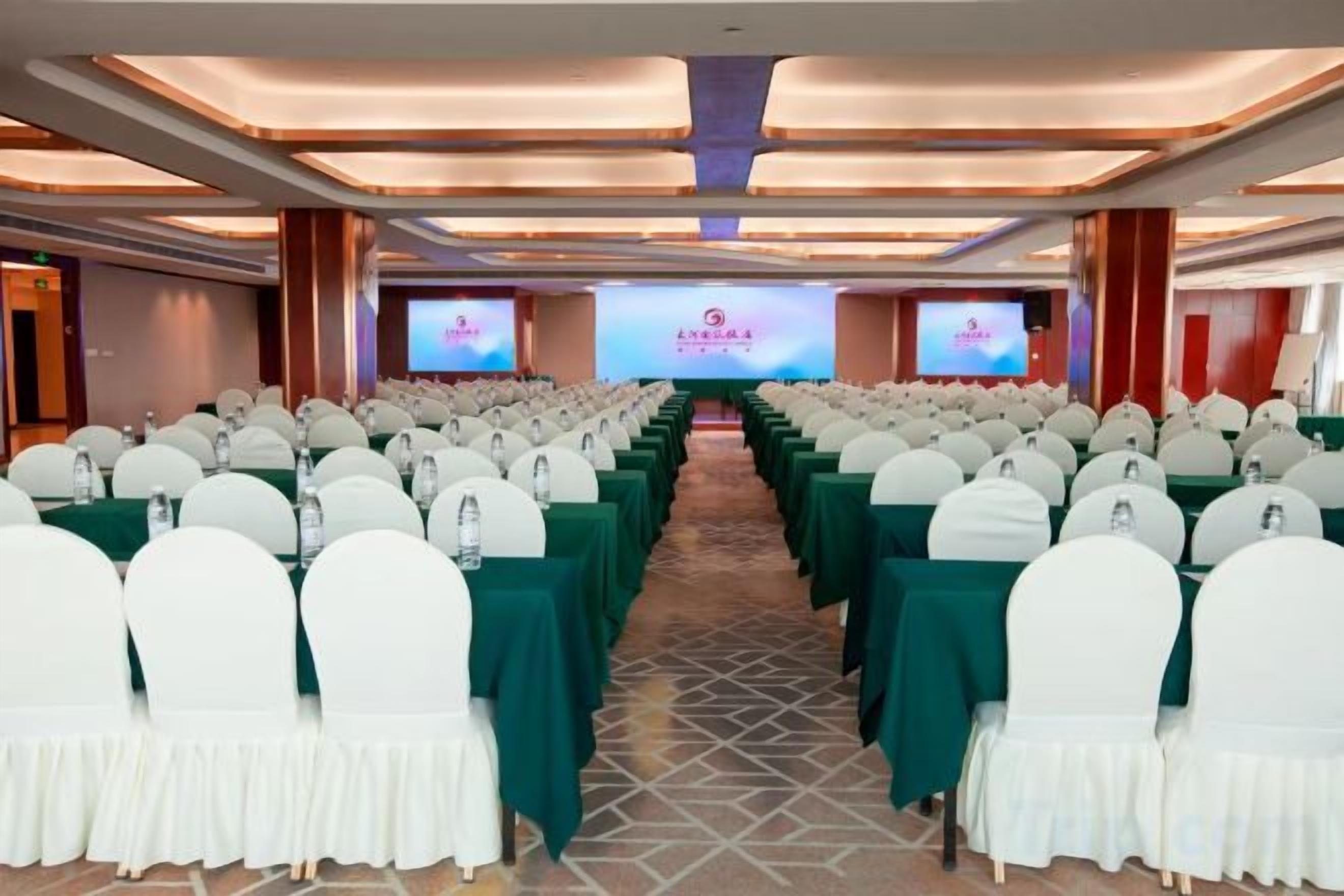 Un salón de conferencias con filas de sillas blancas, manteles verdes y un escenario con una pantalla para presentaciones.