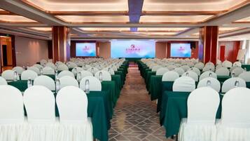 Un salón de conferencias con filas de sillas blancas, manteles verdes y un escenario con una pantalla para presentaciones.