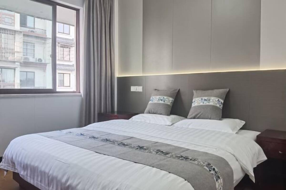 Habitación de hotel con una cama grande, una mesita de noche, una ventana con cortinas y vistas a edificios.