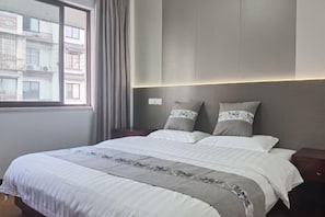 Hotellihuone, jossa on suuri sänky, yöpöytä, ikkuna verhoilla ja näkymä ulkona oleviin rakennuksiin.
