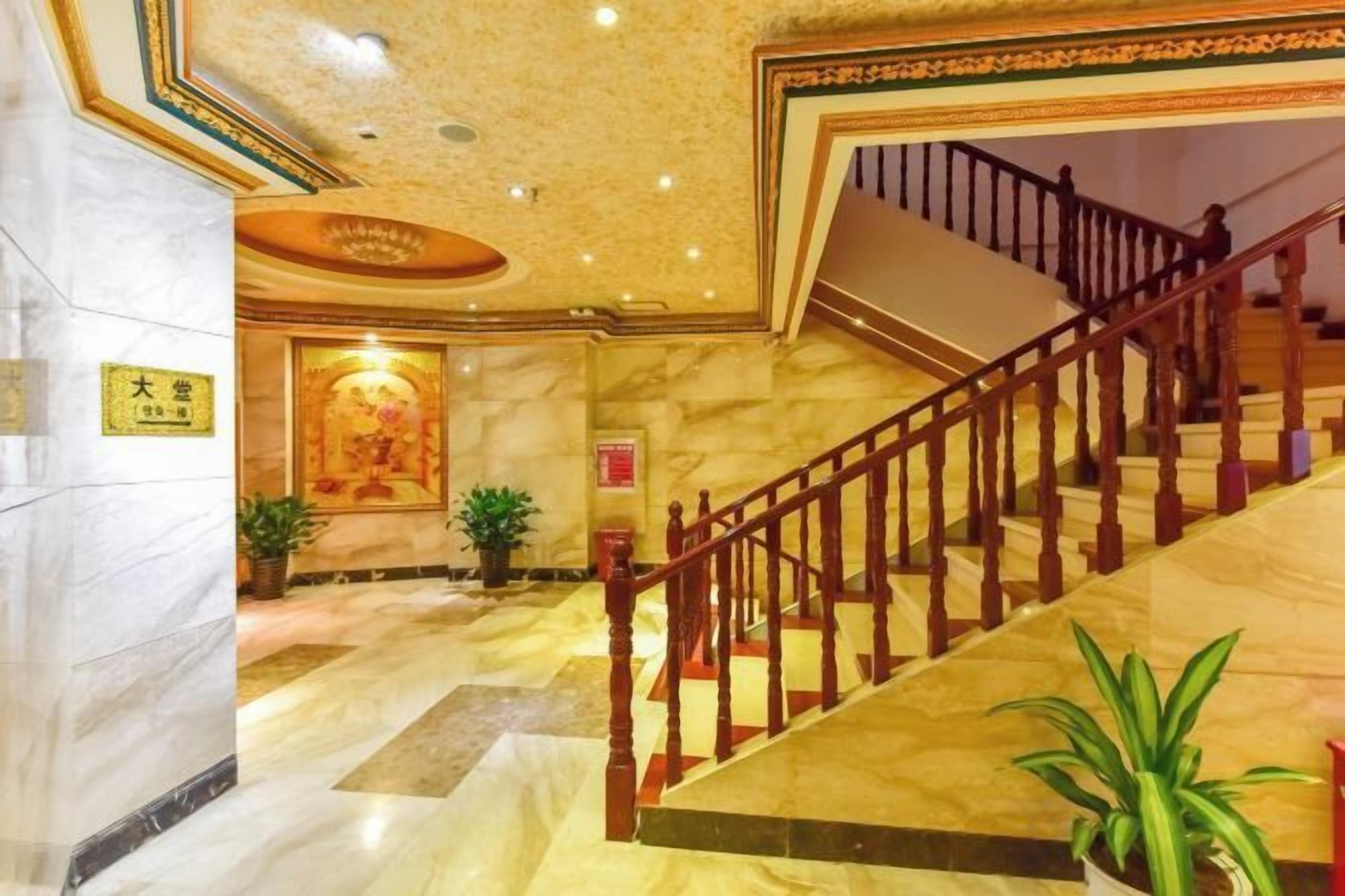 Un vestíbulo de hotel con suelo de mármol, una escalera con barandillas de madera y plantas en macetas.