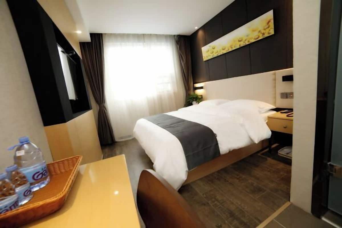 A hotel room with a bed, a desk, a chair, a nightstand, a lamp, a window with curtains, and a wall-mounted artwork.