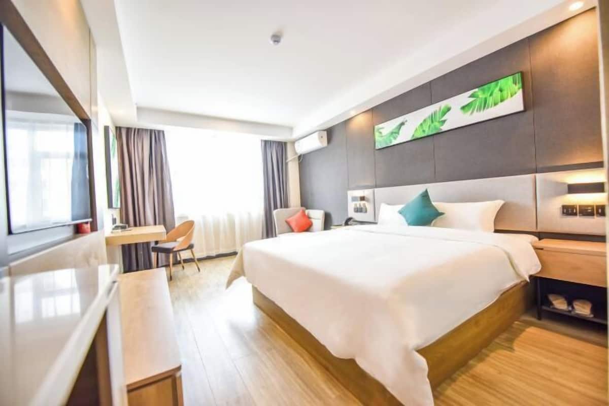 A modern hotel room with a large bed, a desk, and a chair. There is a window with curtains, a wall-mounted artwork, and a bedside table with a lamp.