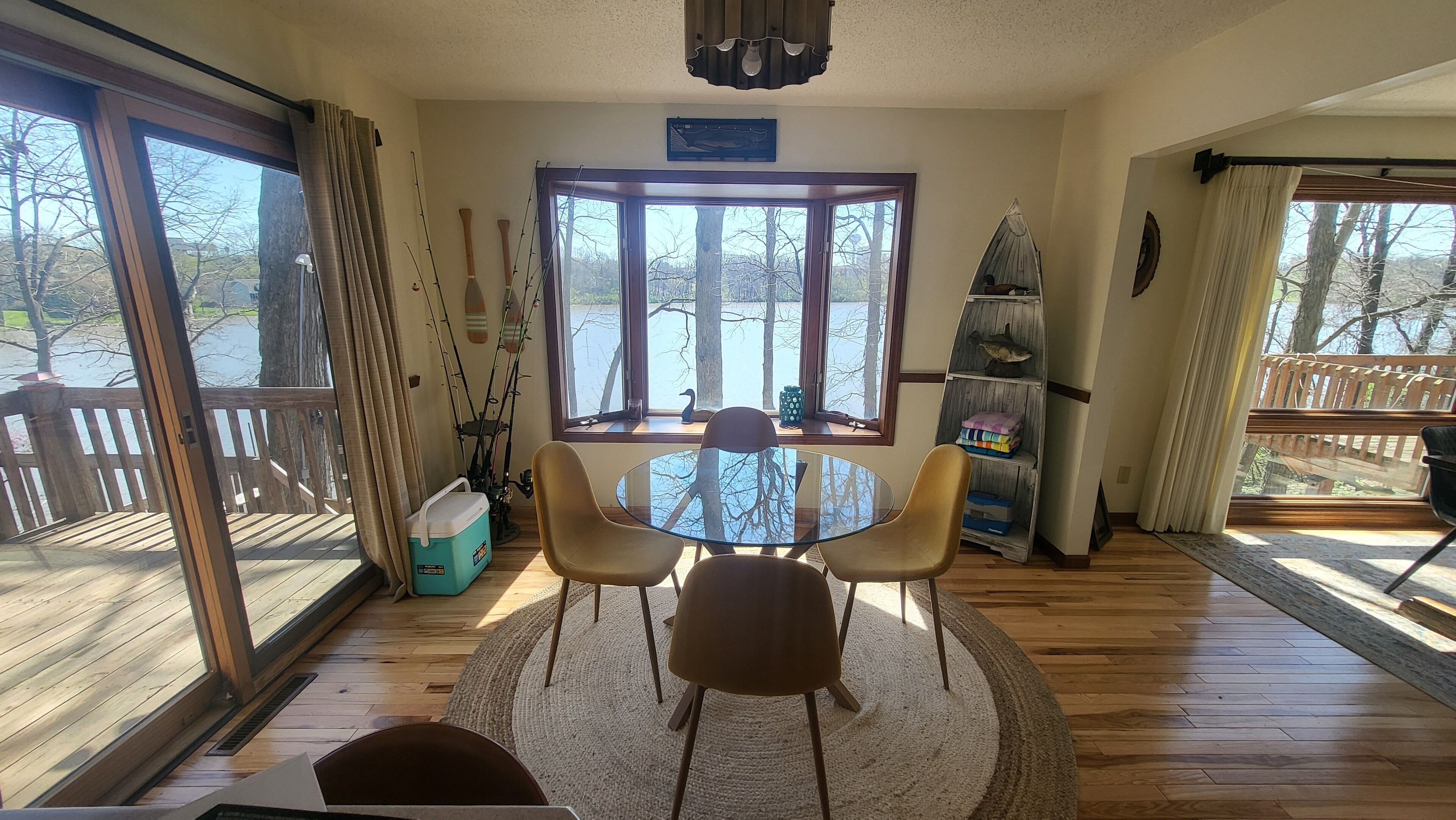 Une pièce avec une table ronde en verre, deux chaises, un petit tapis, une fenêtre donnant sur des arbres et une étagère avec des objets décoratifs.