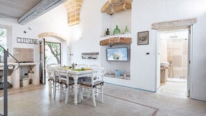 A dining area with a white table and chairs, a kitchen visible through an archway, and a stone fireplace.