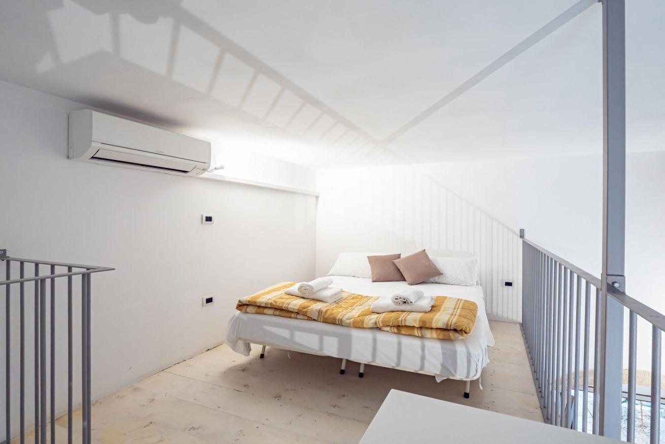 A modern bedroom with a bed, white walls, an air conditioning unit, and a balcony with a railing.
