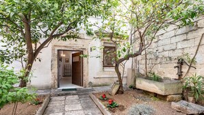 A courtyard with a stone pathway leading to a door, surrounded by plants and a stone bench.