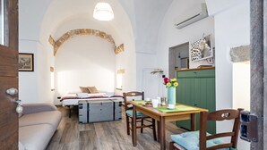 A cozy room with a bed, a dining table with chairs, a small kitchen area, and a stone archway. - GuestHost - Palazzo Rolli - The Room of the Barrels (Galatone)