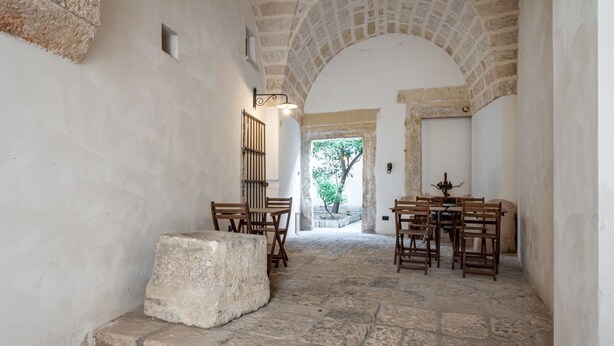 A narrow stone corridor with a stone bench, wooden chairs, and a table. - GuestHost - Palazzo Rolli - The Room of the Barrels (Galatone)