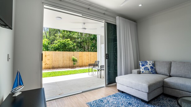 Una sala de estar moderna con un sofá, una mesa de centro y un puerta corrediza de vidrio que da a un patio con cerca de madera y follaje.