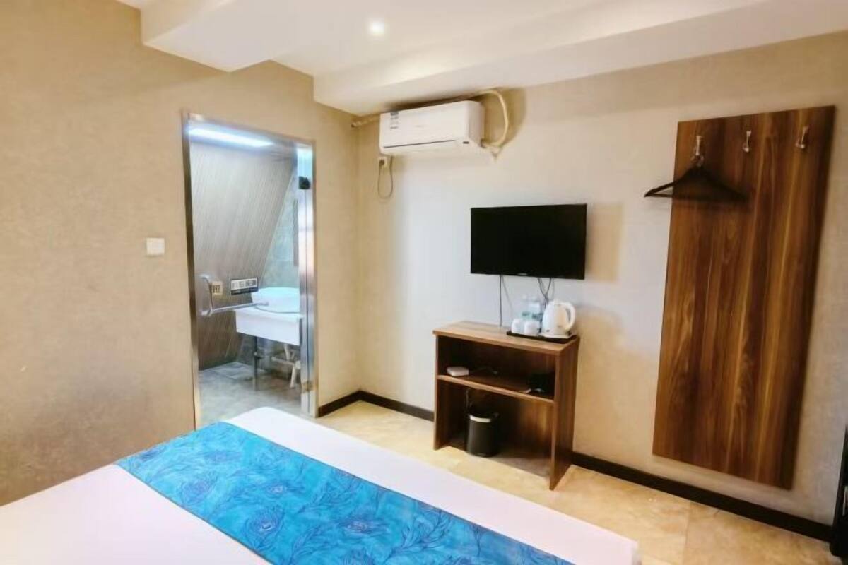 A hotel room with a bed, a television, a wooden wardrobe, a small shelf with toiletries, and an air conditioning unit.