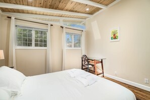 A hotel room with a bed, a desk, and a chair. There are windows with curtains, a painting on the wall, and a wooden ceiling.