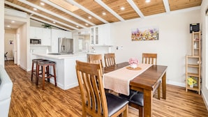 Une cuisine moderne avec des meubles blancs, un coin repas doté d’une table et de chaises en bois, et un plafond orné de poutres apparentes et équipé d’un éclairage encastré.