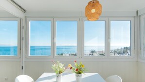 A bright room with a white table, two chairs, and a hanging light fixture. Large windows offer a view of the sea and a distant cityscape. - Magic of the ocean, Front Line Beach Apartment (TORREMOLINOS)