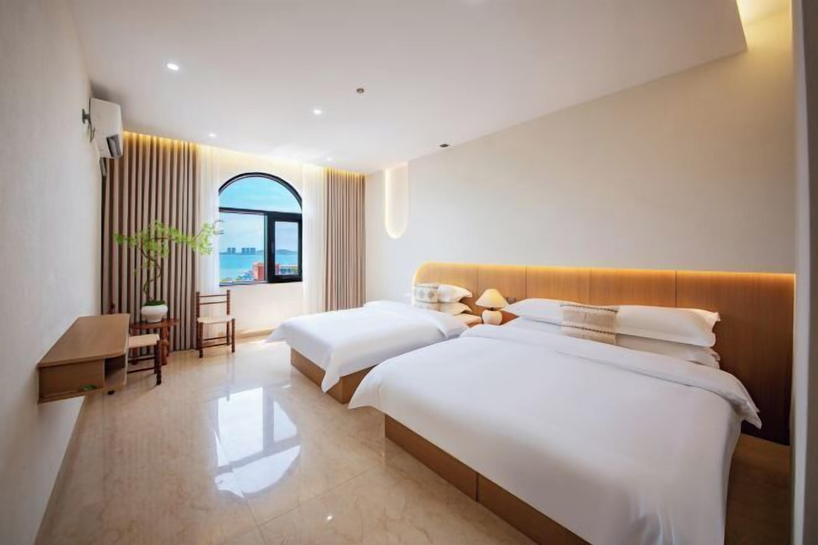 Ein Hotelzimmer mit zwei Betten, einem Schreibtisch, einem Stuhl, einer Zimmerpflanze und Blick auf die Stadt durch ein großes Fenster.