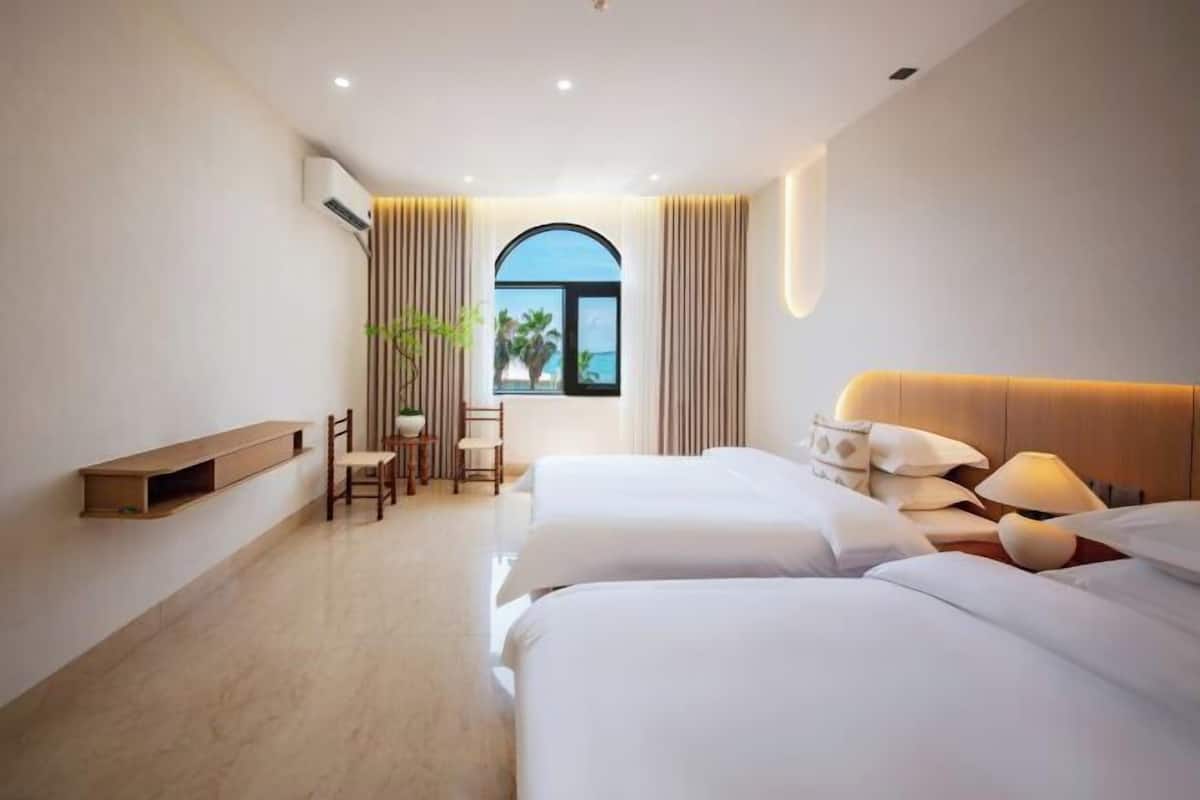 Habitación de hotel con dos camas, un escritorio con sillas, una ventana con cortinas y vista al mar.