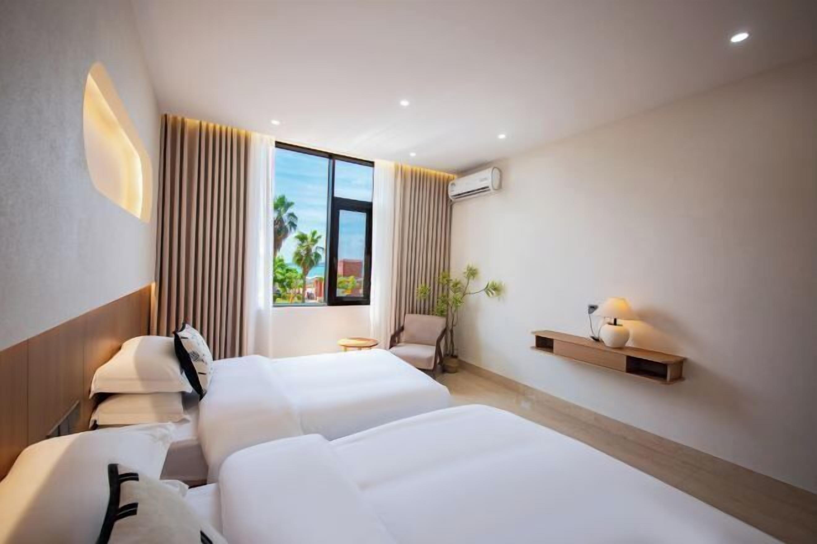 A hotel room with two beds, a bedside table, a chair, a potted plant, a wall-mounted shelf with a lamp, and a window with curtains.