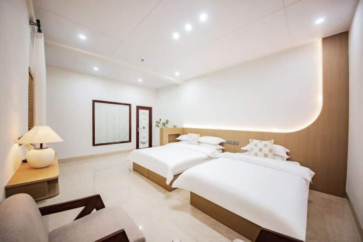 Una habitación de hotel moderna con dos camas, una mesita de noche con lámpara, una silla y una estantería en la pared.