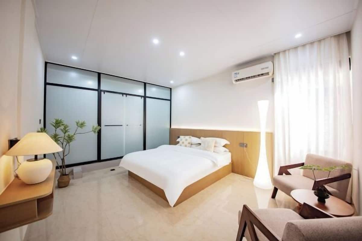 Una habitación de hotel moderna con una cama grande, una mesita de noche con lámpara, una silla y una planta en maceta.