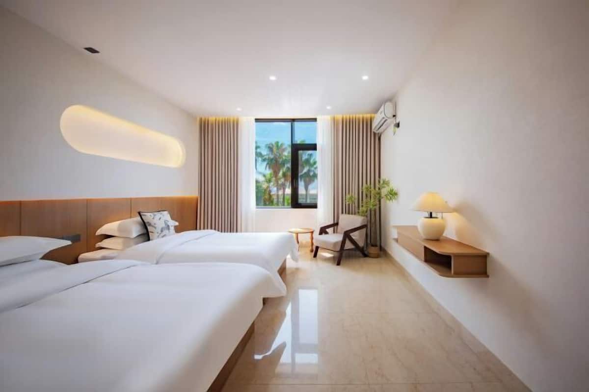 Habitación de hotel con dos camas, un espacio para sentarse, una lámpara y vista a palmeras a través del ventanal.