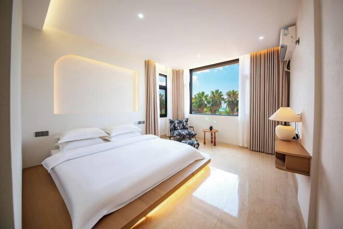 Habitación de hotel moderna con una cama grande, una mesita auxiliar, una silla y una ventana con vistas a palmeras.