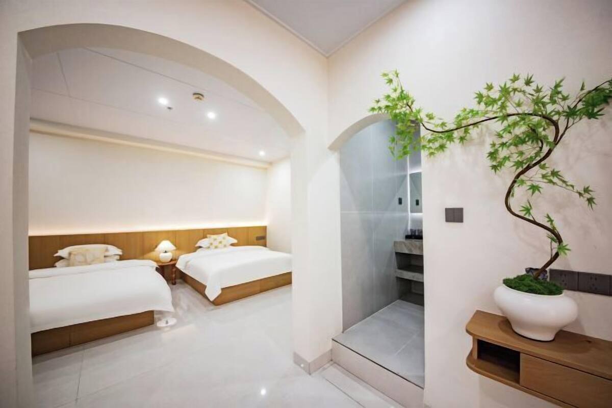 Una habitación de hotel moderna con dos camas, una mesita de noche con lámpara, una planta grande en maceta y un espejo.