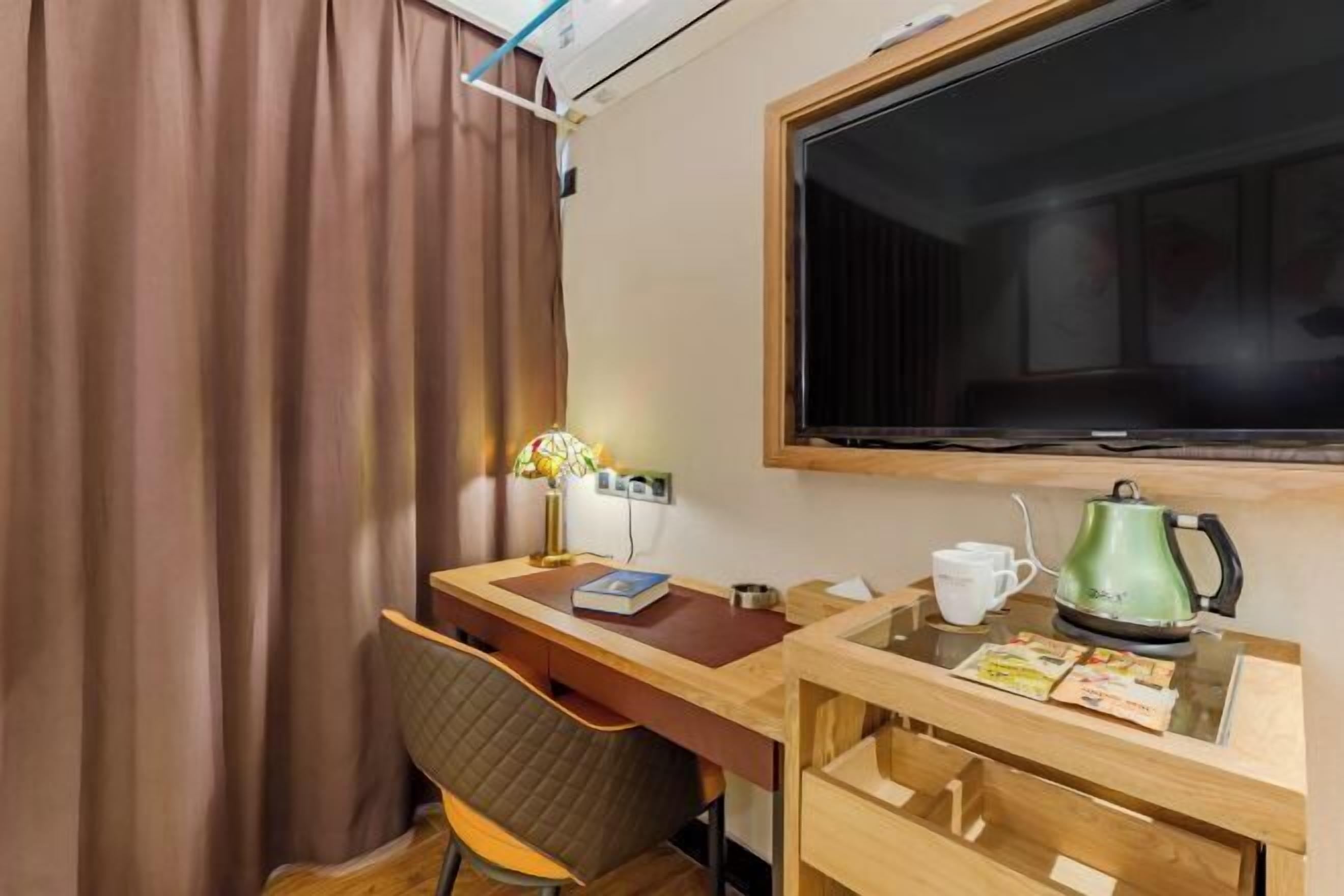 A hotel room with a wooden desk, a chair, a television, a kettle, and a window with curtains.