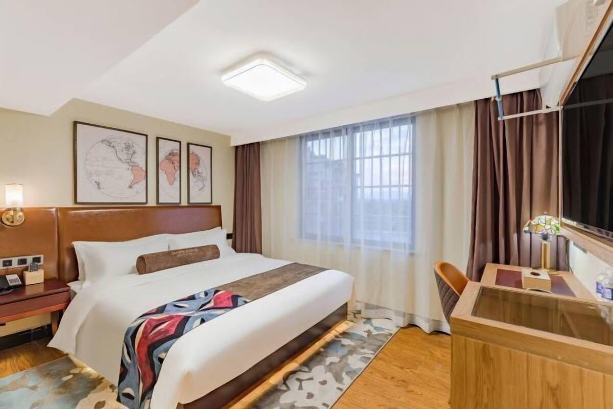 Quarto de hotel com cama, escrivaninha, cadeira, mesa de cabeceira, luminária e uma janela grande com cortinas.