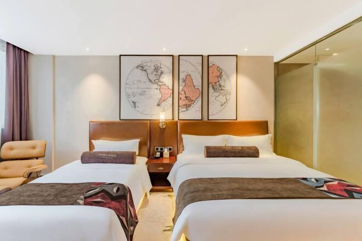 Quarto de hotel com duas camas, uma mesa de cabeceira, uma luminária e uma janela grande.