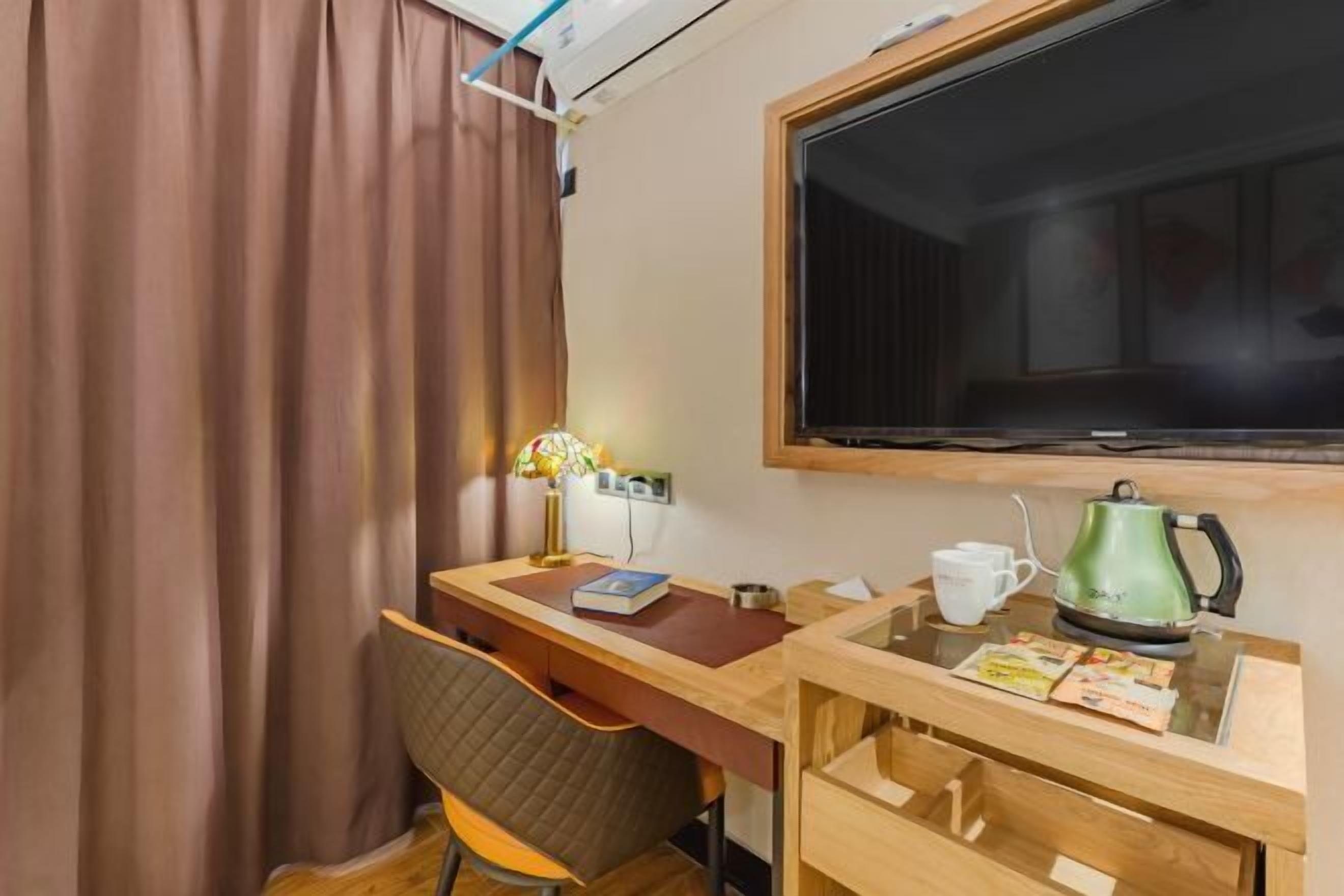 A hotel room with a wooden desk, a chair, a television, a window with curtains, and a kettle on a side table.
