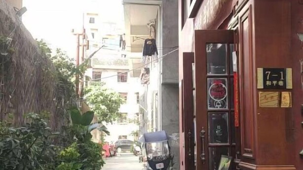 Un carreró estret amb un patinet aparcat, una porta de fusta i un rètol, i un edifici amb balcons.