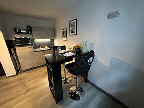 A compact kitchen with a microwave, coffee maker, and sink. A dining area with a black chair and table set for two, featuring wine glasses and plates.