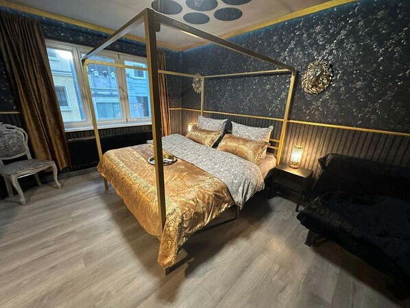 A four-poster bed with a gold canopy, a bedside table with a lamp, a dark sofa, and a vintage chair in a room with a dark, textured wall and a window with curtains.