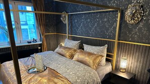 A four-poster bed with a gold and white patterned bedspread, a bedside table with a lamp, and a window with curtains.