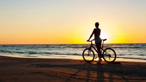 A person standing on a beach with a bicycle, facing the ocean at sunset.