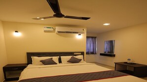 A hotel room with a large bed, a ceiling fan, an air conditioning unit, a bedside table, a nightstand, and a window with curtains. - Sri Srinivasa Residency (Puducherry)