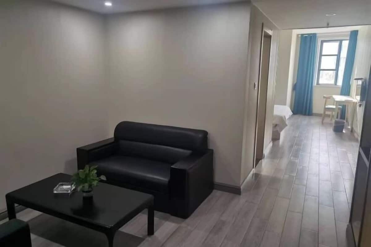 A modern living room with a black leather sofa, a small table with a plant, and a hallway leading to a bedroom with a bed and a chair.