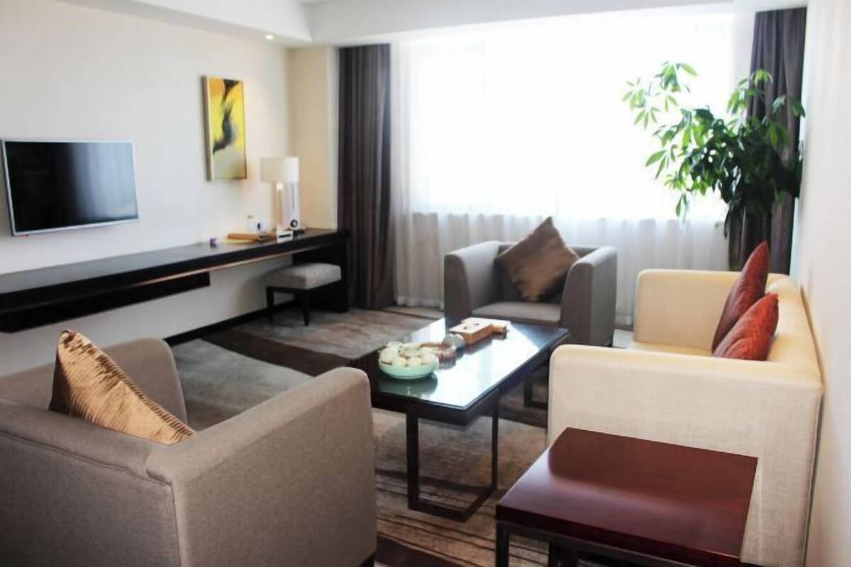 A modern hotel room with a flat-screen TV, a sofa, a chair, a coffee table, and a large window with curtains.