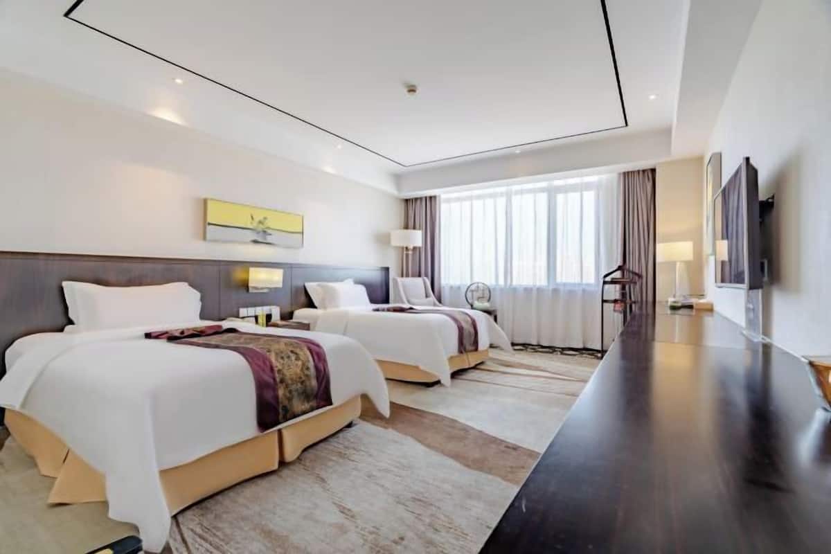 A modern hotel room with two beds, a long dark wooden table, and a large window with curtains.