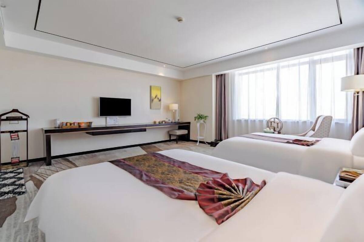 A hotel room with two beds, a flat-screen TV, a desk, and a large window with curtains.