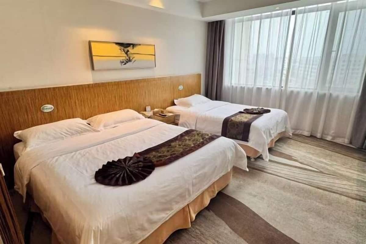 A hotel room with two beds, a wooden headboard, a wall-mounted lamp, and a large window with curtains.