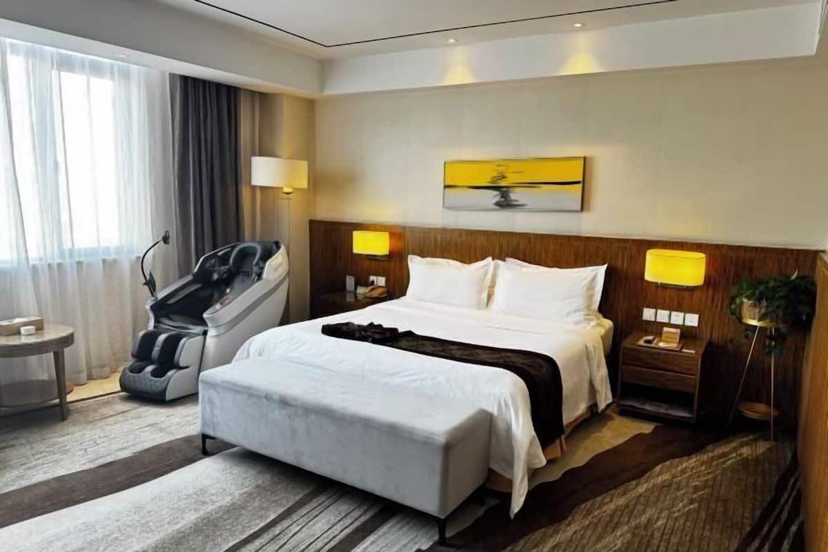 A hotel room with a bed, bedside lamps, a massage chair, a small table, and a wall-mounted artwork.