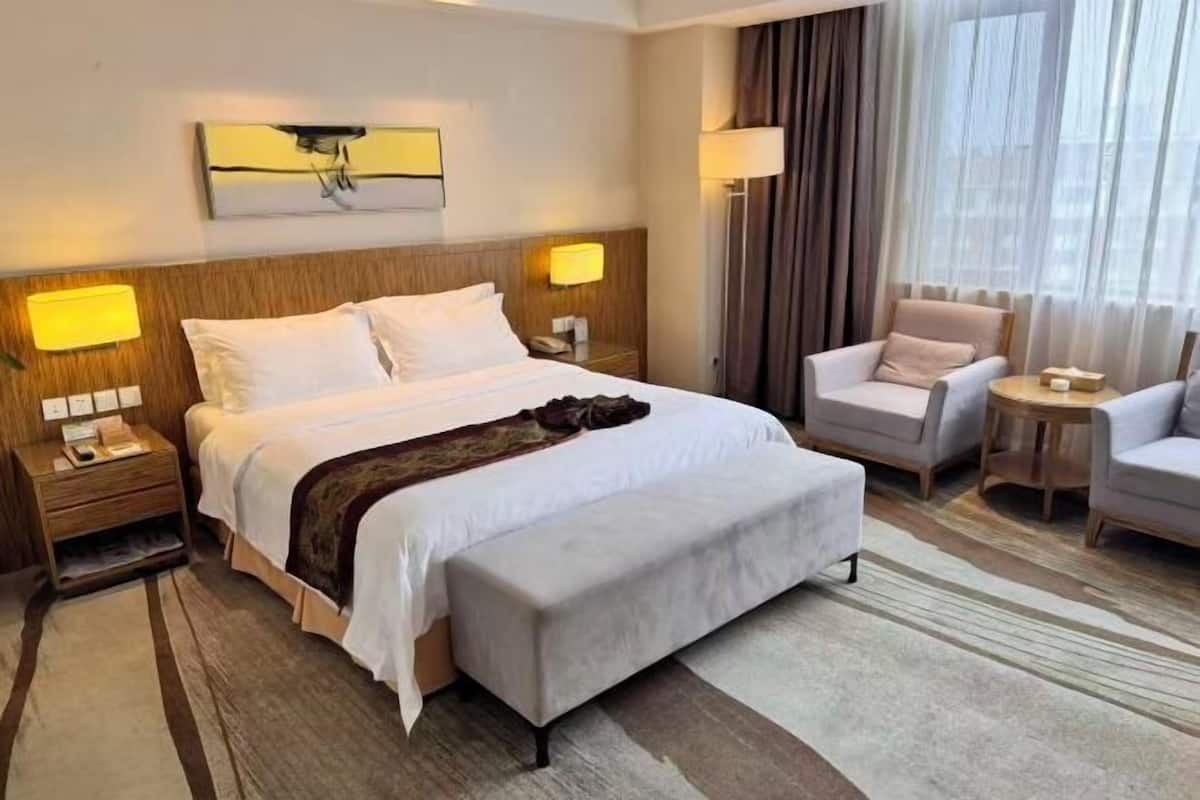 A hotel room with a bed, two bedside tables, a chair, a small table, and a large window with curtains.