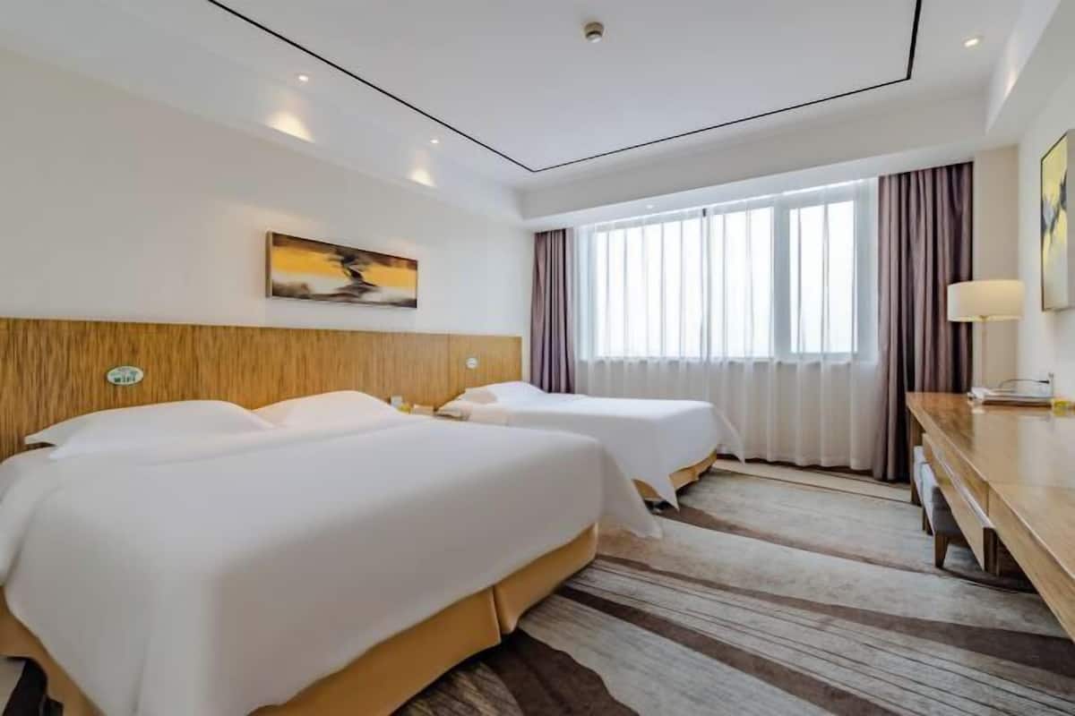 A hotel room with two beds, a wooden headboard, a large window with curtains, a wooden desk, and a painting on the wall.