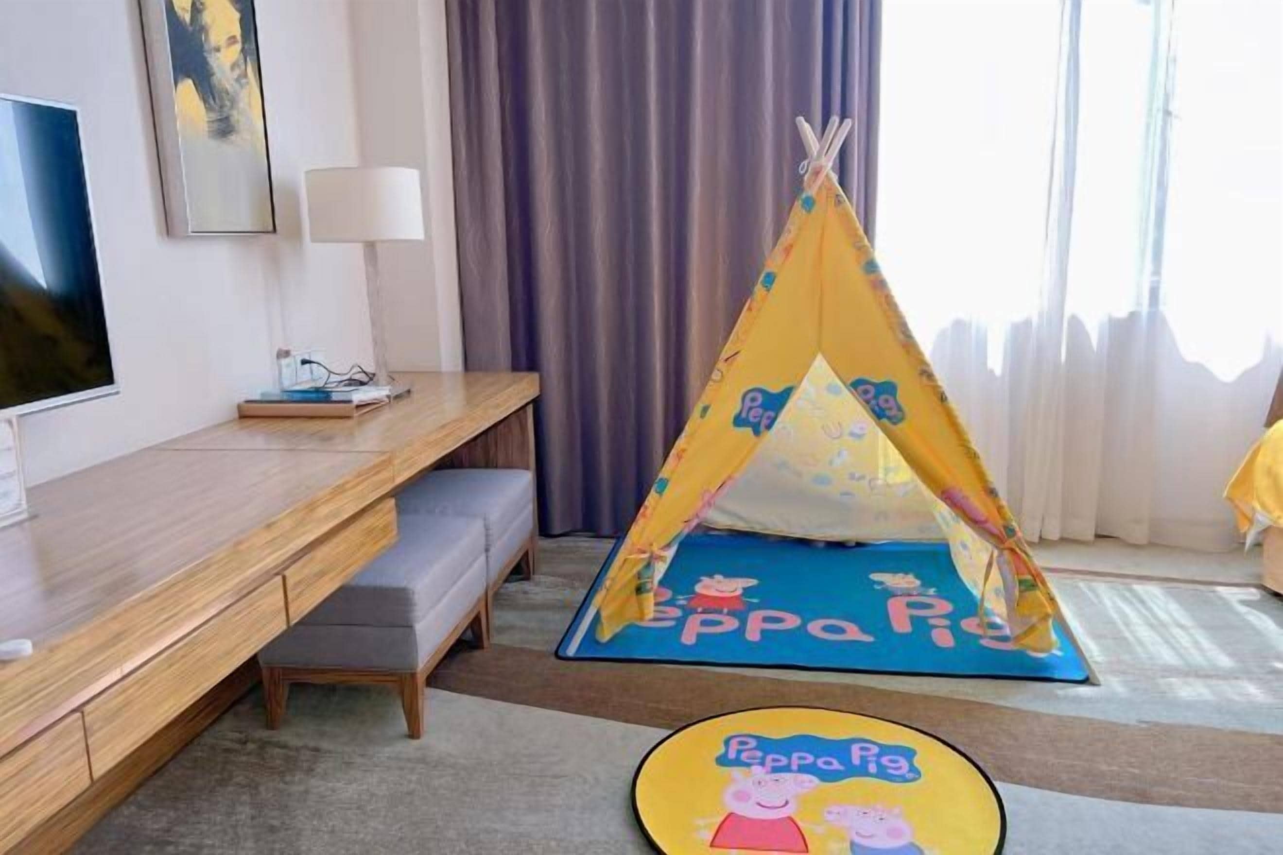 A hotel room with a wooden desk, a grey bench, a Peppa Pig-themed play tent, and a Peppa Pig round mat.
